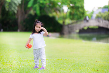Obraz premium Happy girl playing small orange ball in green grass with family. Picnic with parents on holiday. Winter in central Thailand. Cool weather. Cute 3 year old child, sweet smile, Child scratching eyebrows