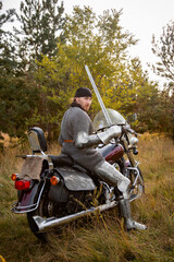 A medieval warrior in armor sits on a motorcycle with a sword in his hands on the background of the forest. Knight biker concept.