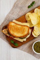 Homemade Pesto Chicken Sandwich with Potato Chips on a rustic wooden board on a white wooden surface, top view. Flat lay, from above, overhead.