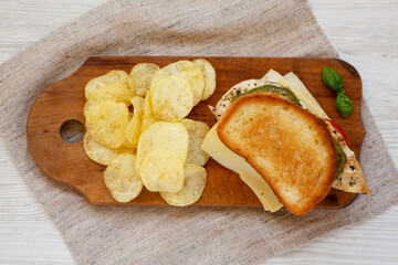 Homemade Pesto Chicken Sandwich with Potato Chips on a rustic wooden board on a white wooden background, top view. Flat lay, from above, overhead.