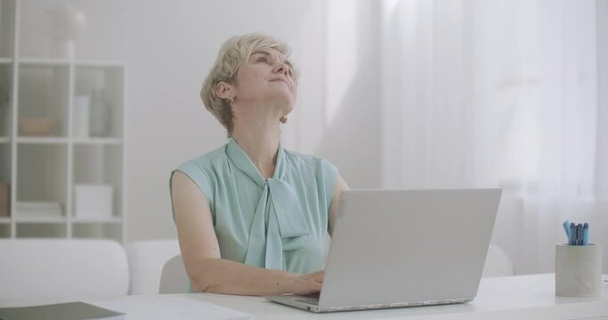 tired aged woman is stretching hands and shoulders during work with laptop, sitting at table and typing messages