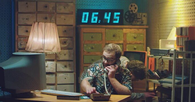 Two Caucasian males programists working in retro style room. Man in glasses answering vintage wired telephone ringing and talking at computer monitor. Men nerds of 80's or 90's. Hackers of 70's.