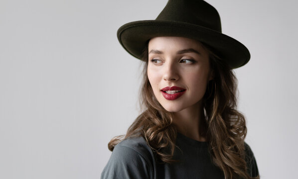 Close up portrait of  female in stylish black hat with brown curls and wine red lips posing in studio isolated on gray background. Autumn fashion concept.