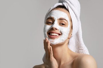 Smiling pretty girl in towel applying eco-friendly skincare mask for acne on face isolated on gray...
