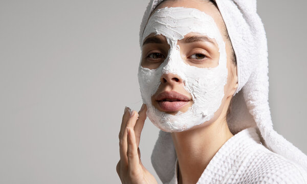 Young woman in a towel applying facial white mask at her face at home, isolated on a gray background. Skin care concept. Advertising poster of natural cosmetics
