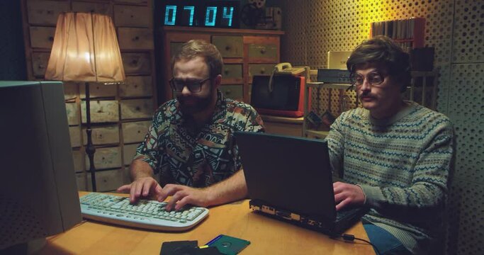 Caucasian males nerds sitting at table, talking and working on computers. Two retro young men programists typing on keyboards, studying and mocking. Vintage style of 90's. Friends hackers of 80's.