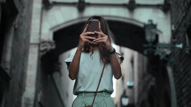 Beautiful long-haired brunette making photo under the arch. Young attractive woman taking pictures of stunning buildings. Pretty tourist makes selfie in a beautiful city. Girl with mobile phone.