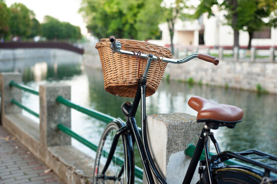 Rear view of a city women's bicycle with a wicker brown basket. Bicycle leaning against the wall of the river bank in the urban part of the city. The river in the background