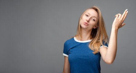Funny young blonde female shoots in temple, dressed in casual blue t shirt, isolated on gray...