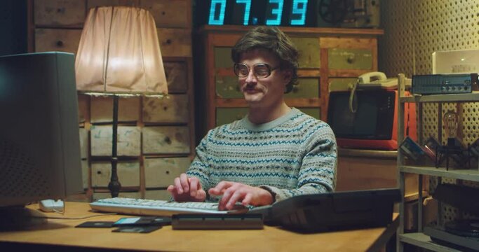 Caucasian male nerd in glasses with mustache sitting at desk in retro room, working and caressing computer. Man programist typing on keyboard while studying. Vintage style of 90's. Hacker from 80's.