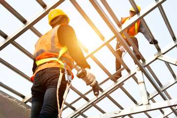 Construction workers wear safety straps while working on the building's roof structure at a construction site. Roofer using a pneumatic nail gun.
