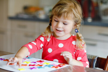 Little toddler girl playing with different colorful stickers and painting flowers. Concept of activity of children during pandemic corona virus quarantine. Child learning colors with parents at home