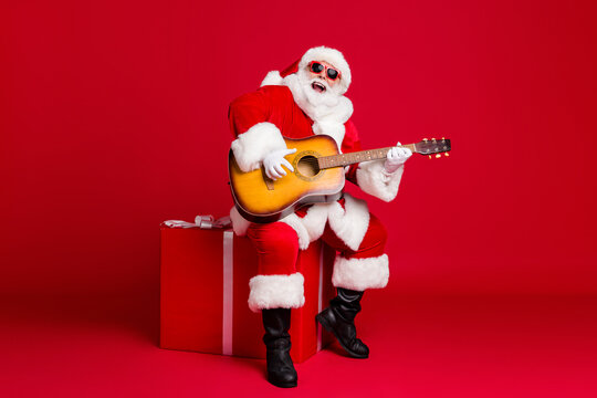 Full length photo of retired old man white beard hold guitar sit box play sing christmas carol song wear santa x-mas costume sunglass headwear leather boot isolated red color background