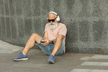 Attractive and smiling bearded old senior man with white hair enjoying music from smart phone outdoor wearing white headphones.