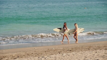 Long shot footage of two slim young adult Caucasian women walking along picturesque ocean coast holding surfboards