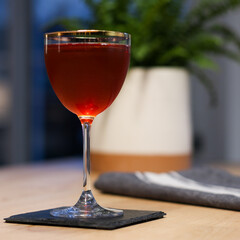 Elysian Cocktail in Nick & Nora gold rimmed glass on wood table with plant in background with a deep red color