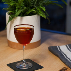 Elysian Cocktail in Nick & Nora gold rimmed glass on wood table with plant in background with a deep red color