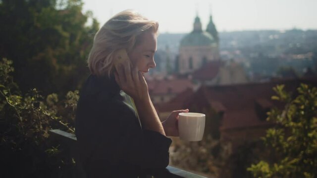 Businesswoman On The Balcony Laughing When Talking On The Phone While Drinking A Cup Of Hot Coffee. - medium shot