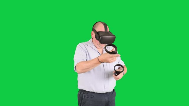 Caucasian male, mid 50s wearing light blue shirt interacts in immersive virtual reality with handheld controllers & VR headset. Innovative emerging technology devices/goggles. Greenscreen, mid shot.