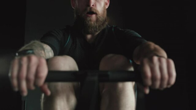 Big strong man with tattoos and beard trains on the rowing machine in slow motion.