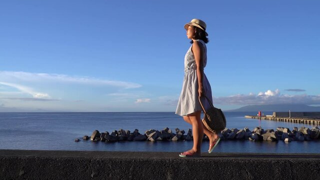 Japanese girl in summer dress holding bag walking in front of Ocean on beautiful clear day - wide sideways tracking shot