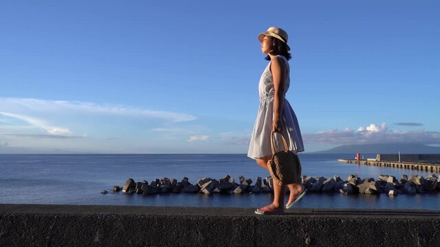 Japanese girl in summer dress holding bag walking in front of Ocean on beautiful clear day - wide sideways tracking shot SLOW MOTION