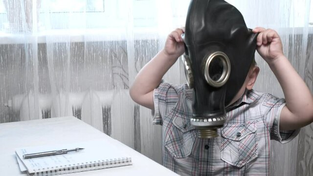 little short-haired caucasian boy in checkered shirt sitting at table at home. pen and notebook in front of him. he tries to wear gas mask, his father helps him. protection during covid-19 quarantine