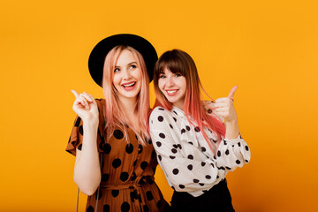 Indoor positive portrait of two gorgeous girls posing over yellow background. Vintage outfit, black hat, candid smile. Party mood.