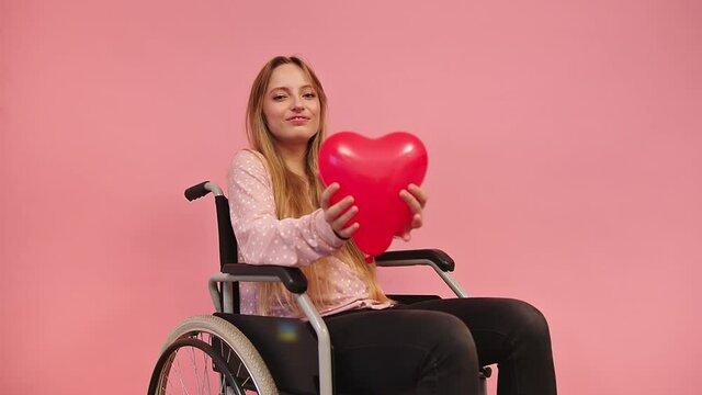 Profile of Young Smiling Female Sitting in Wheelchair With Heart Shaped Balloon. Health Care for Disabled People Concept, Static Full Frame Isolated on Pink Background