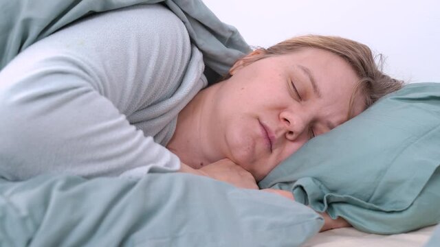 Young blonde tired woman in pajamas is sleeping, mumbling and snoring in bed under warm blanket at home. Health and breathing problems, somnambulism.