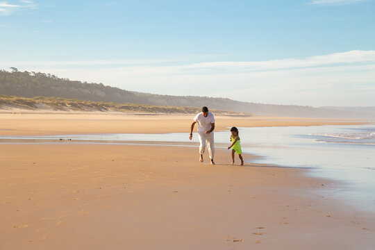 Dad And Little Girl Running On Wet Golden Sand On Beach. Front View, Wide Shot. Family Outdoor Activities Or Leisure Time By Sea Concept