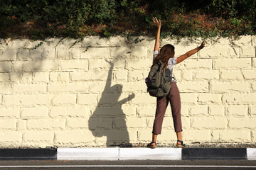 Young woman tourist with backpack stands with her back to the road at the edge of the curb and waving her arms, looking at her shadow on light stone wall