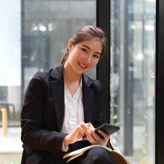 Female in suit using smartphone while relaxed sitting in glass wall meeting room