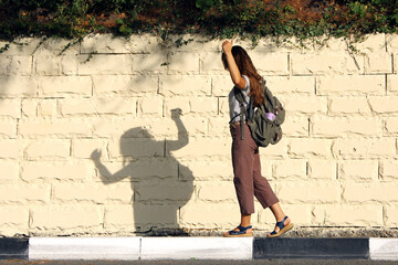 Young woman tourist with backpack behind her back has fun, playing with shadow, waiting for transport at the edge of the road