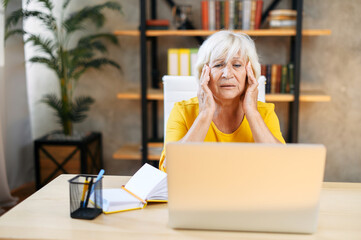 Tired senior business woman is using laptop for work indoor. A beautiful older lady has headache from overworking