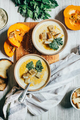 two plates of pumpkin soup next to greens pumpkin and bread croutons on a white wooden table top view close up