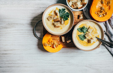 two plates of pumpkin soup next to greens pumpkin and bread croutons on a white wooden table top view close up
