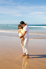 Handsome young dad spending leisure time with little daughter on beach at sea, holding kid in arms. Side view. Parenting and outdoor activities concept