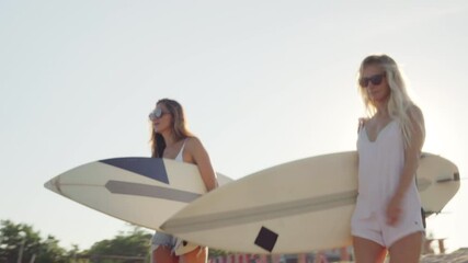 Two cheerful Caucasian women with tanned skin wearing light clothing going to ocean beach with surfboards