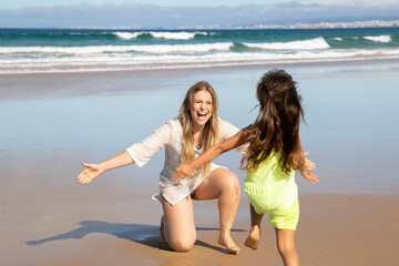 Fototapeta premium Happy excited mom opening arms for catching and hugging little daughter. Girl running to mother on beach at sea. Parenting and outdoor activities concept