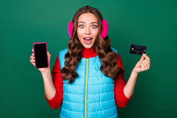 Photo of pretty cute young lady smiling showing credit card telephone shocked look online shopping sale discounts wear pink ear warmers blue vest red sweater isolated green color background