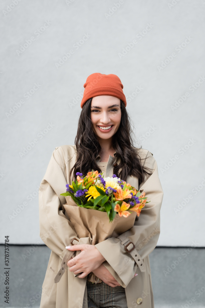 Sticker stylish woman in beanie hat and trench coat holding bouquet of flowers near grey wall