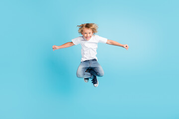 Fototapeta na wymiar Photo of small child jump funny look camera raise hands wear white shirt jeans sneakers isolated blue color background