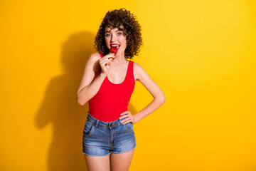 Photo of lovely pretty excited young lady hand hips hold strawberry put open mouth eating keep healthy diet wear red singlet unclothed shoulders isolated bright yellow color background