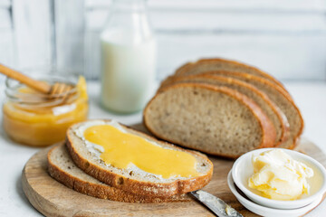 Slice of sourdough bread with honey, butter and milk on wooden chopping board, white table. Healthy, organic country breakfast with natural morning light. Copy space. Freshly baked bread.