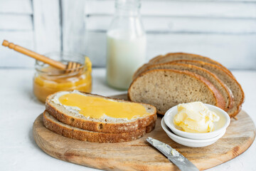 Slice of sourdough bread with honey, butter and milk on wooden chopping board, white table....