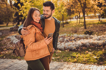 Photo of positive two people students go in yellow autumn park guy embrace girl use smartphone wear backpack coat