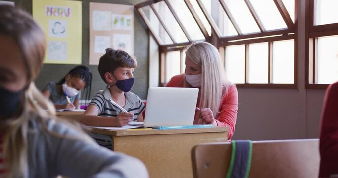 Female teacher and boy wearing face mask using laptop in class at school