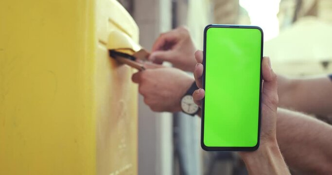 Close up view of female hand holding smartphone with mock up screen. Unknown person hands putting letter in post box at background. Concept of greenscreen and chroma key. Outdoors.