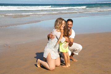 Happy parents hugging little daughter on wet sand at sea. Mom, dad and kid enjoying leisure time on beach at ocean. Parenting and childhood concept